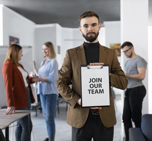 A man stands in an office, holding a clipboard that says Join Our Team