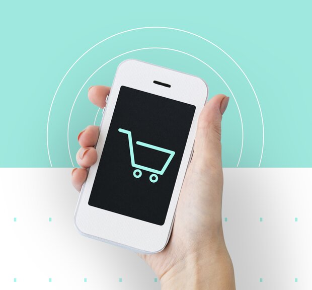Eine Hand hält ein Smartphone, auf dessen Bildschirm ein Symbol für einen Einkaufswagen angezeigt wird, das Zahlung durch PAYONE hinweist.